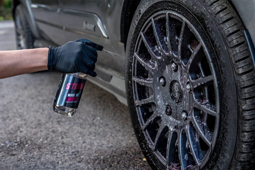Black-gloved hand spraying wheel cleaner onto a dark car rim